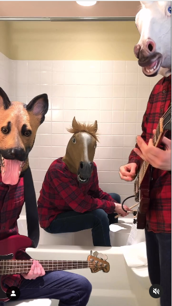 A still from a video with three people wearing flannel shirts. A man wearing a horse mask is sitting in a bath tub with a bucket for a drunk. A man in a dog mask is sitting on a toilet and playing bass. A man in a unicorn mask is playing guitar.