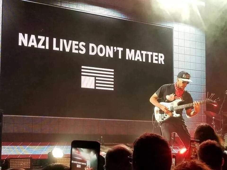 Tom Morello playing on stage in front of a sign that says "NAZI LIVES DON'T MATTER" with an upside-down American flag under the word
