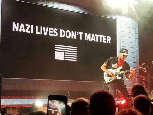 Tom Morello playing on stage in front of a sign that says "NAZI LIVES DON'T MATTER" with an upside-down American flag under the word